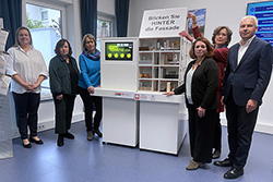 Ein Foto aus der Ausstellung mit dem Modell eines Wohnhauses zur Verfügung. Es zeigt v. l. n. r. die Leiterin der Polizei im MTK Carina Lerch, Andrea Bartels-Pipo von „Frauen helfen Frauen“, Erste Stadträtin Renate Mohr, Petra Gokkenbach von „Frauen helfe